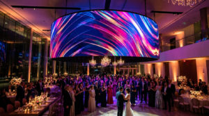 Un couple danse au centre d'un mariage élégant, sous un grand écran LED incurvé affichant des motifs lumineux et colorés. Les invités applaudissent.