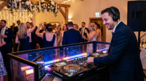 Un DJ souriant avec des écouteurs mixe sur une console Pioneer lors d'un mariage animé, avec des invités dansant en arrière-plan.