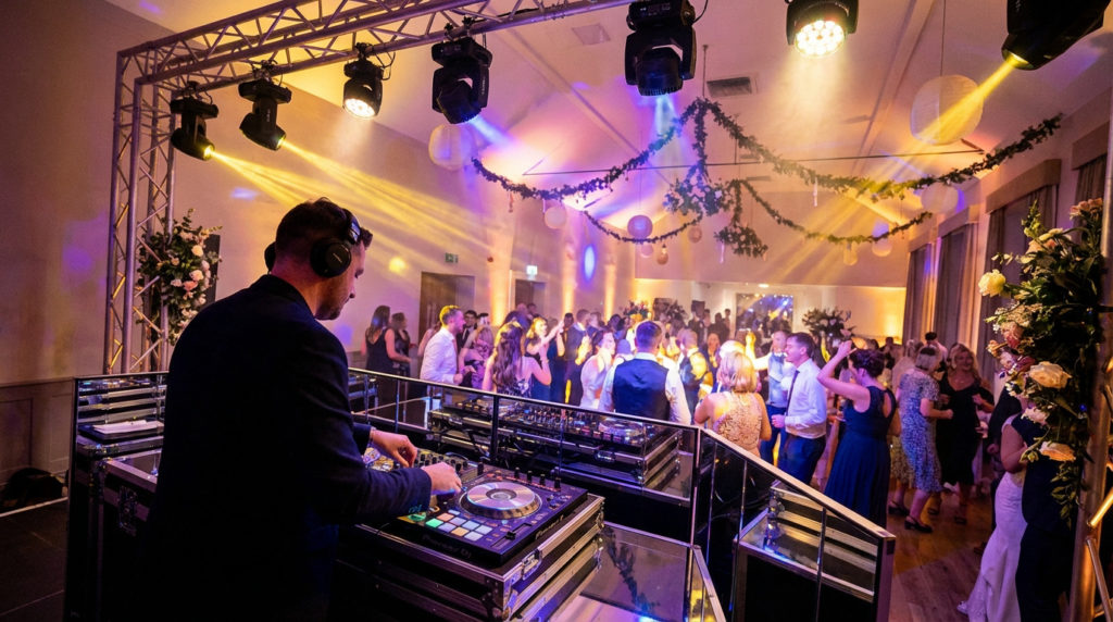 Un DJ avec un casque aux platines, mixant pour des invités de mariage dansant dans une salle de réception festive.