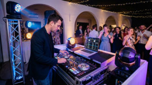 DJ homme mixant avec console et ordinateur portable à une soirée festive. Invités souriants dansent en arrière-plan sous des lumières.