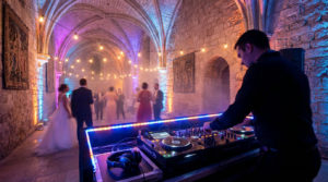 DJ mixant lors d'un mariage dans une salle médiévale aux voûtes en pierre, ambiance lumineuse violette et bleue avec guirlandes.